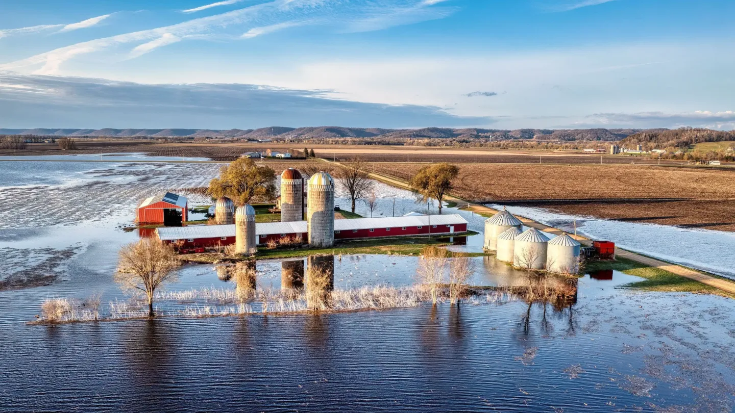 Aerial view of a farmstead with grain silos surrounded by floodwater across low-lying fields.