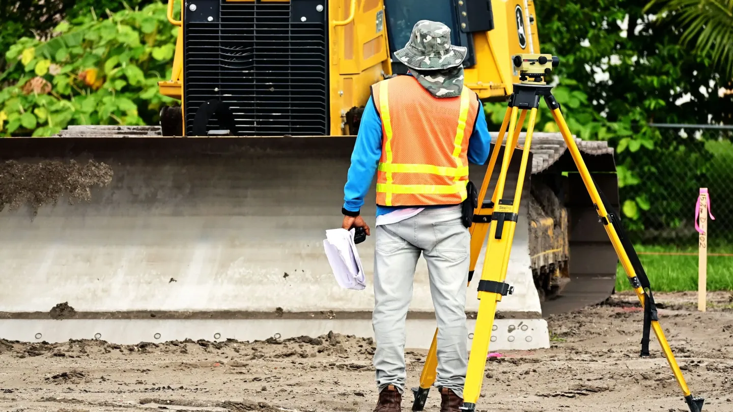 A surveyor in a high-visibility vest sets up a tripod on freshly graded ground in front of a bulldozer.